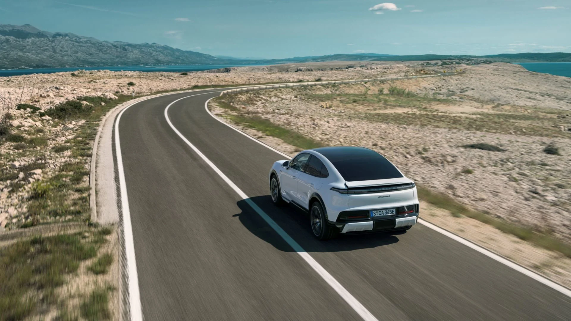 Rear view of the 2026 Porsche Cayenne Turbo Electric driving on a rural road at sunset, showing its full-width LED light bar and sporty stance.